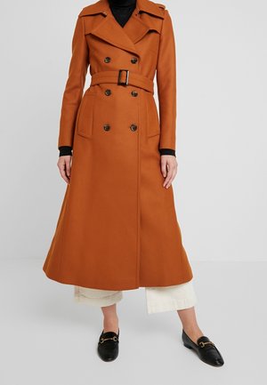 Kobieta w długim, dwurzędowym płaszczu trencz w kolorze spalonym pomarańczem, z paskiem, w zestawieniu z białymi, krótkimi spodniami i czarnymi mokasynami.