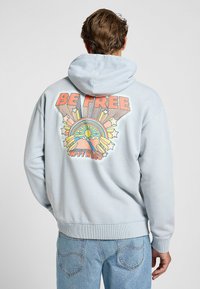 Felpa leggera blu chiaro con un vivace design grafico sul retro, completo di testo "BE FREE", motivi di sole, arcobaleno e stelle. Materiale in misto cotone.