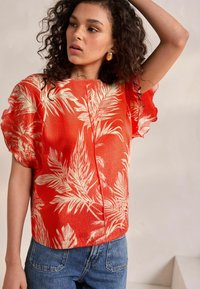 Short-sleeved, textured top in bright orange, featuring white tropical leaf patterns, styled with a relaxed fit and a round neckline.