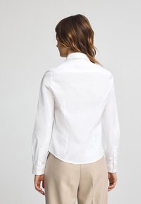 Camisa blanca de manga larga con cuello estructurado, textura suave y diseño ajustado, que presenta puños abotonados y un dobladillo curvado.