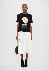 Tricou negru cu un grafic de ou prăjit și text, asortat cu o fustă albă care are detalii din panglică texturată. Geantă neagră și tocuri.