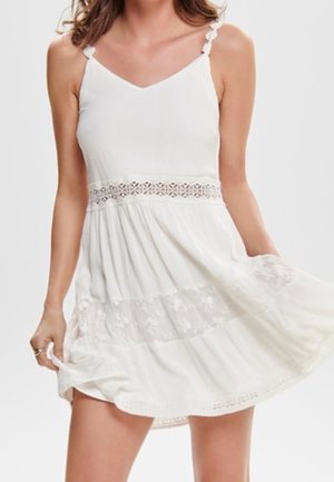 Bez rękawów biała sukienka z koronkowym wykończeniem i haftem kwiatowym, trzymana lekko na zewnątrz przez dłonie kobiety na wysokości bioder.