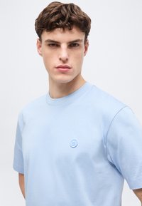 Νεαρός με κοντά καστανά μαλλιά που φοράει γαλάζιο μπλουζάκι με στρογγυλή λαιμόκοψη και μικρό κυκλικό λογότυπο στο στήθος.