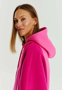 Světlý růžový mikina s měkkou texturou, s prostornou kapucí, volným střihem a detailem na šňůrku. Hladký povrch, bez viditelných vzorů.