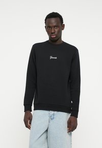 Felpa nera con maniche lunghe e scollo rotondo, con ricamo bianco "Yosemite" sul petto. Indossata con jeans azzurri chiari.