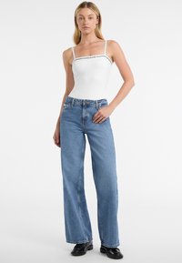 Biela priliehavá body s tenkými ramienkami, čierny text loga, v kombinácii s modrými nohavicami s vysokým pásom a širokými nohavicami a čiernymi robustnými topánkami.