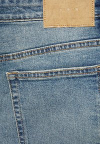 Gros plan sur un jean en denim bleu montrant une poche arrière avec des coutures orange et une étiquette de marque en cuir beige cousue.