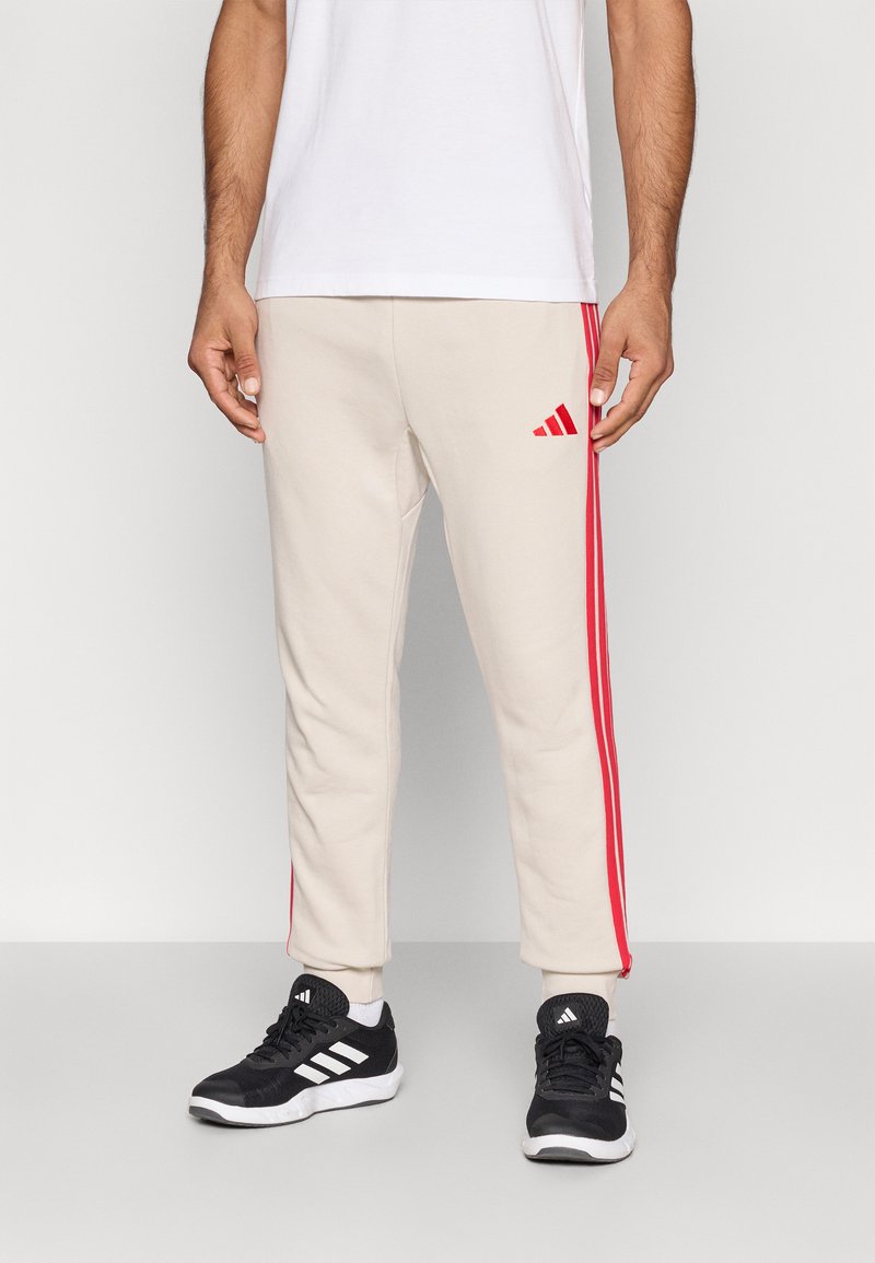 Smėliniai treningai su raudonomis šoninėmis juostomis, su siuvinėtu logotipu. Dėvimi su balta marškinėliais ir juodais sportiniais batais.