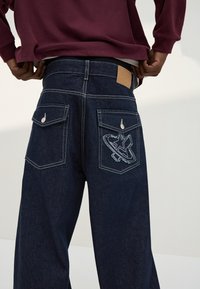 Persoon met donkerblauwe wijde jeans met klepzakken aan de achterkant, waarvan één zak een gestikte ster- en ringontwerp heeft, en een bordeauxrode trui.