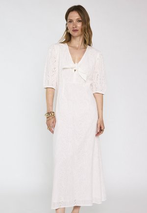 Vrouw draagt een witte eyelet midi-jurk met pofmouwen en een strik bij de hals, gecombineerd met gouden armbanden en ringen.
