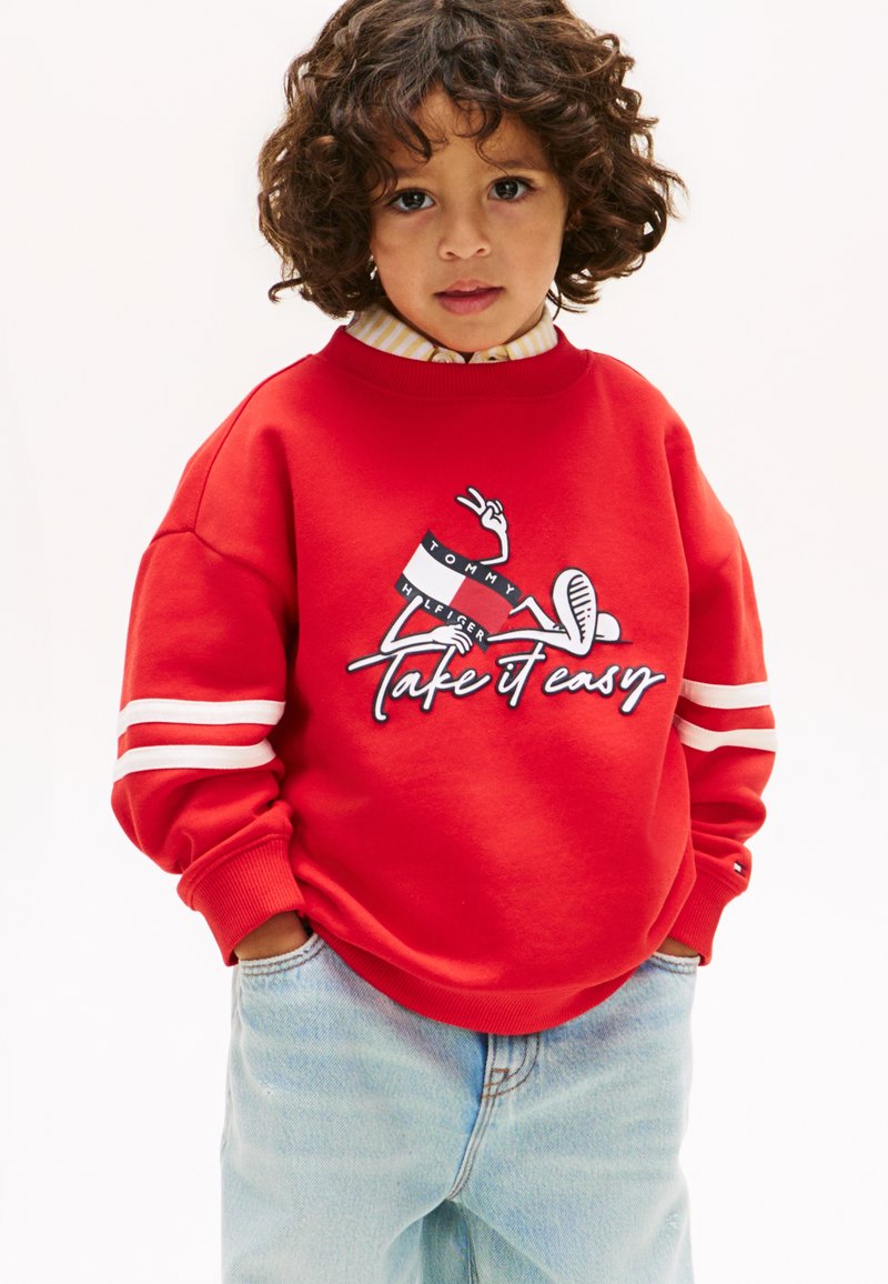 Παιδί με σγουρά μαλλιά που φοράει κόκκινο φούτερ με λευκές ρίγες και το κείμενο «Take it easy», συνδυασμένο με ανοιχτό μπλε τζιν και χέρια στις τσέπες.