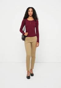 Haut henley à manches longues bordeaux avec trois boutons, associé à un pantalon beige ajusté et des ballerines noires, ainsi qu'un petit sac à épaule noir.