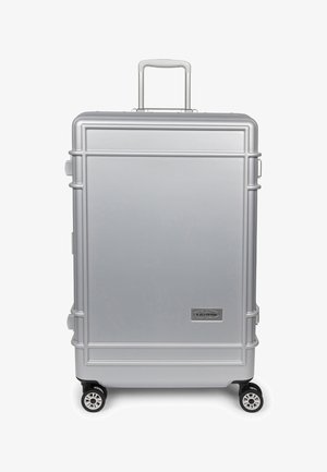 Maleta rígida plateada con asa extensible, cuatro ruedas giratorias y un pequeño logo rectangular de la marca en la esquina inferior derecha del frontal.
