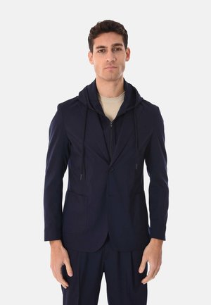 Uomo che indossa un blazer blu navy con cappuccio attaccato e pantaloni abbinati, in piedi davanti a uno sfondo bianco uniforme.