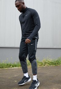 Atletyczny mężczyzna w czarnym dresie Nike i sportowych butach, stojący na wybrukowanej nawierzchni przy szarej ścianie z zielonymi roślinami w tle.