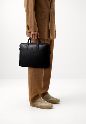 Serviette en cuir noir avec doubles poignées, surfaces lisses et texturées, arborant un logo centré et une fermeture éclair, tenue par une personne portant un pantalon beige.