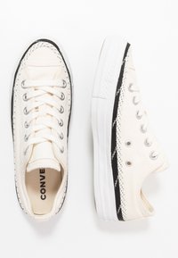 Baskets basses en toile blanche avec lacets blancs, détails noirs sur la semelle et œillets en métal, vues de dessus et de côté.