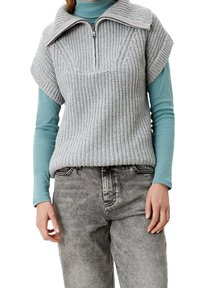 Ljusgrå stickad tröja med hög krage, korta ärmar och dragkedja framtill, lager på lager över en turkosa långärmad topp och grå jeans.