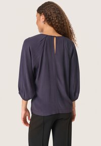 Blusa blu navy con maniche a 3/4, dettaglio plissettato sul retro e apertura a forma di chiave. Tessuto morbido e strutturato, abbinata a pantaloni neri.