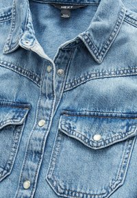 Chemise en denim bleu clair avec un col pointu, deux poches poitrine et des boutons-pression en argent. Présente des coutures doubles et une texture douce.