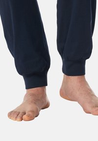 Pantalones de chándal de algodón en color azul marino con puños elásticos, que ofrecen un corte relajado. Pies descalzos con uñas visibles y un ligero vello en las piernas.