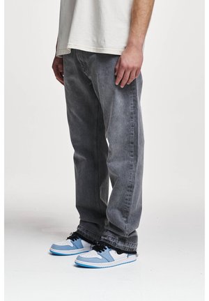 Mand iført falmede sorte jeans med frynsete kanter og hvide sneakers med lyseblå detaljer står på en hvid baggrund.