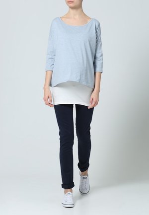 Blusa de manguita larga color azul claro, holgada, superpuesta a una camisa blanca, combinada con pantalones oscuros de corte slim y zapatillas blancas.