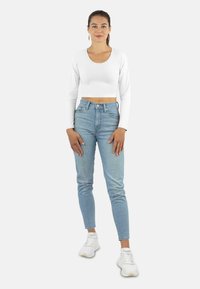 Weißes, langärmeliges Cropped-Top mit rundem Halsausschnitt, kombiniert mit hochgeschnittenen hellblauen Jeans und weißen Sneakers. Schlichter Stil mit minimalen Details.