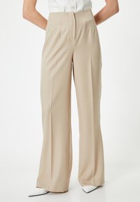 Beige hoge taille broek met een wijd model, voorzien van een gladde textuur en geplooide details aan de voorkant. Gestyld met puntige witte schoenen.