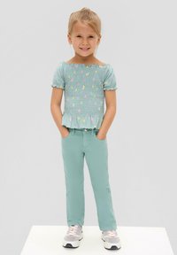 Jasnozielona bluzka z marszczeniami i kwiatowymi wzorami, zestawiona z dopasowanymi zielonymi spodniami. Dziewczynka stoi pewnie, z dłońmi w kieszeniach, w tenisówkach.