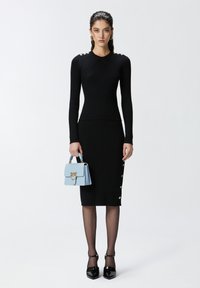 Abito lungo a maniche lunghe nero a coste con dettagli in bottoni e taglio aderente. Il modello tiene una borsa azzurra chiaro con accessori dorati. Tacchi neri a punta e collant.