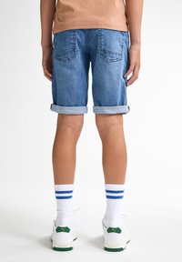 Shorts de mezclilla en azul claro, con los puños doblados, y dos bolsillos traseros. Llevados con calcetas blancas con rayas azules y zapatillas blancas con acentos verdes.