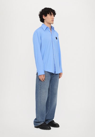 Helles blaues Langarmhemd mit einem schwarzen Herz-Logo auf der Brust, kombiniert mit hellblauen weit geschnittenen Jeans und schwarzen Schuhen.