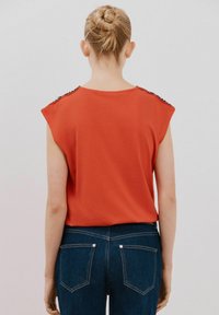 Rød ærmeløs top med skulderdetaljer, parret med mørkeblå jeans. Stoffet ser blødt ud med en glat tekstur.