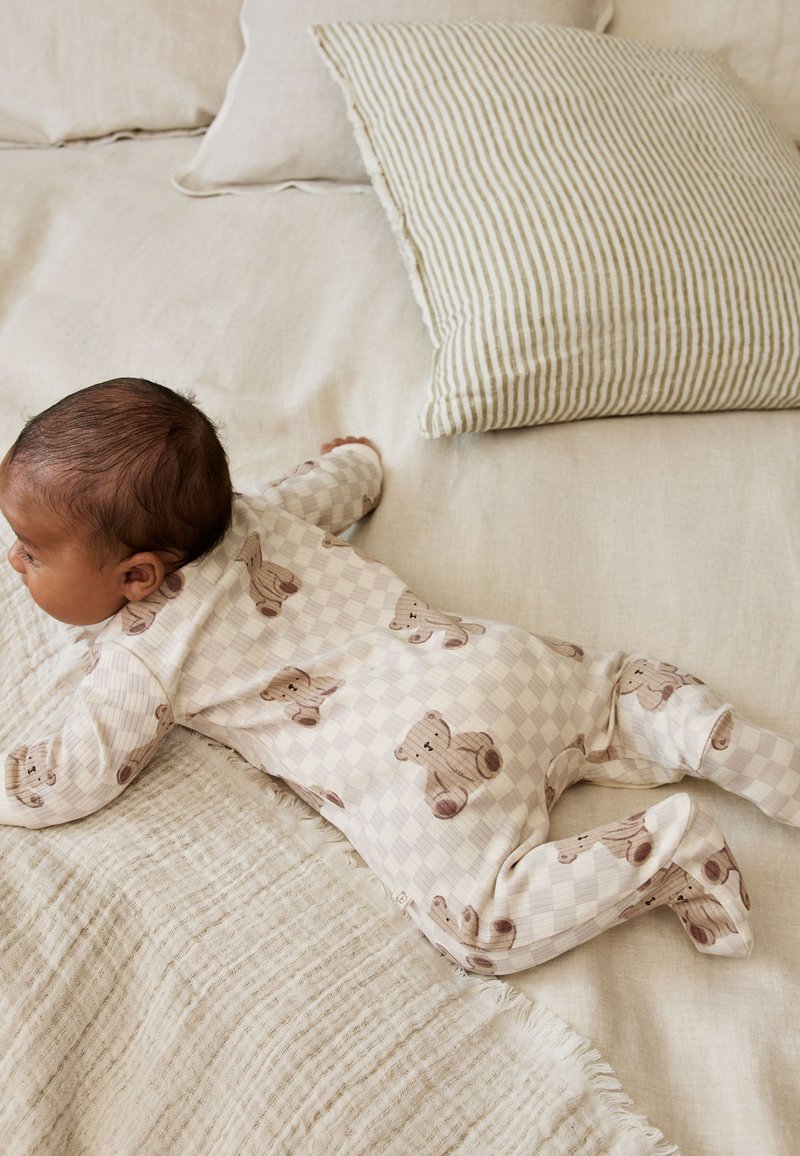 Bebé tumbado boca abajo en la cama con un pijama estampado con ositos de peluche, junto a una manta crema texturizada y una almohada a rayas.
