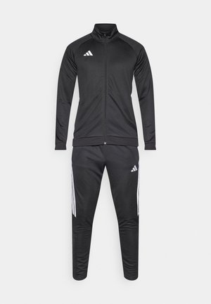 Fekete, sima anyagból készült melegítő; cipzáras kabáttal magas gallérral és fekete nadrággal, fehér átlós csíkokkal.