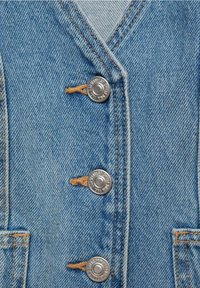 Veste en jean de couleur bleu clair, ornée de cinq boutons-pression argentés, de coutures marron et d'un col en V. Tissu texturé avec un détail de poche visible.