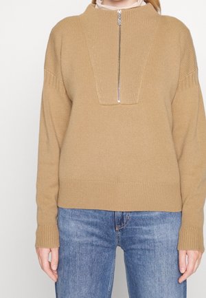 Beige striktrøje med foran lynlåsdetaile, lange ærmer og ribstrikkede ærmekanter, båret med blå denim jeans.