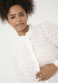 Blusa blanca con cuello volante, que presenta un sutil patrón de zigzag y cierre de botones. Tejido ligero y transparente con mangas largas.