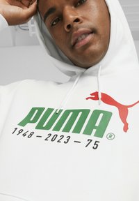 Sudadera blanca con el logo de PUMA en verde y rojo, y el texto "1948 - 2023 - 75" en negro. Capucha con cordones, textura de tela suave.