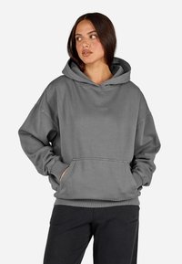 Γυναίκα που φοράει μια φαρδιά γκρίζα κουκούλα με μπροστινή τσέπη και μαύρο παντελόνι, κοιτώντας αριστερά σε μια απλή φόντο.