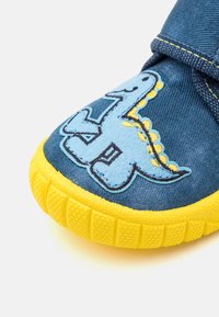 Zapato infantil azul con suela amarilla, que presenta un parche de dinosaurio sonriente en la punta y un cierre de tira de Velcro.