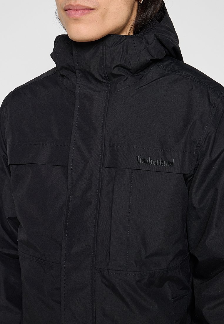 Chaqueta negra impermeable con un alto cuello, cierre con cremallera y logo de Timberland en el pecho. Textura suave con un diseño minimalista.
