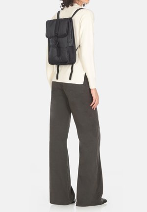 Zaino in nylon nero con chiusura a ribasso, spallacci regolabili e finitura testurizzata, indossato sulla schiena sopra un maglione crema e pantaloni grigi.