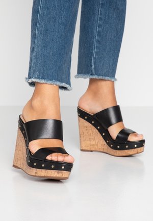 Czarne skórzane sandały na koturnie z korkową platformą, z dwoma szerokimi paskami i metalowymi ćwiekami wzdłuż podstawy. Wysokość obcasa jest znacząca.