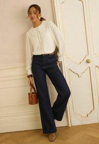 Nő fehér csipkegalléros blúzban, sötét, bő szárú farmerral, világosbarna csizmában, barna bőr vödrötáskával a kezében, falnak dőlve beltéren.