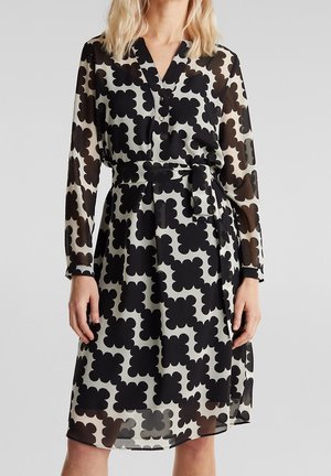 Femme portant une robe noire et blanche mi-longue avec des motifs floraux abstraits audacieux et une ceinture à la taille, dotée de manches longues transparentes.