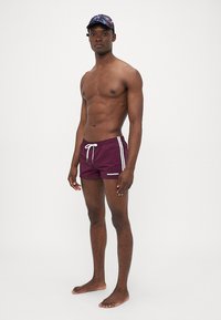 Burgunderfarbene Badehosen mit weißen Seitenstreifen und verstellbarem Bund, präsentiert auf einem shirtlosen Model, barfuß vor einem neutralen Hintergrund.