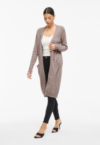 Kvinne som har på seg en lang mauve cardigan over en hvit tanktop, svarte skinny jeans og svarte åpne høyhælte mule-sandaler, som går fremover.