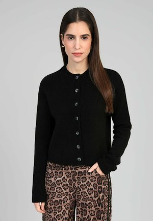 Femme aux cheveux longs et foncés, portant un cardigan noir boutonné et un pantalon marron à imprimé léopard, debout devant un fond gris clair uni.